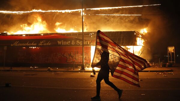 Un manifestante porta la bandera de EEUU boca abajo como símbolo de peligro extremo que vive el país frente un edificio en llamas en Minneapolis (estado de Minnesota), durante las protestas causadas por la muerte de George Floyd, el 28 de mayo del 2020 - Sputnik Mundo