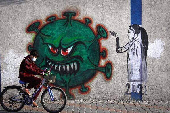 Un joven palestino con una mascarilla puesta monta en bicicleta cerca de una pintura mural que representa a una enfermera que inyecta una vacuna contra el COVID-19 en la ciudad de Gaza. - Sputnik Mundo