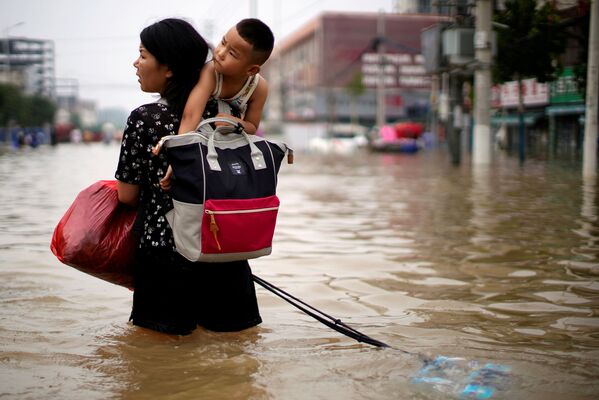 Una mujer con un niño y sus pertenencias recorre una calle inundada en Zhengzhou. - Sputnik Mundo