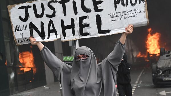 Протестующая держит плакат с надписью Справедливость для Нахеля на фоне горящих автомобилей в конце марша памяти в честь подростка, застреленного полицейским, в парижском пригороде Нантер - Sputnik Mundo