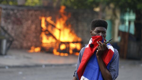 Un hombre, con la cara cubierta por una bandera haitiana, pasa junto al puesto en llamas de un vendedor de arte, incendiado por los manifestantes, 2019 - Sputnik Mundo