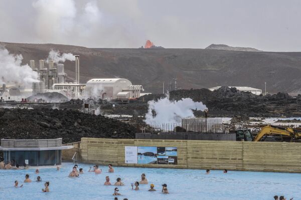 Gente se baña en el popular complejo geotérmico Blue Lagoon con el telón de fondo de un cráter volcánico que escupe lava, en Grindavík, Islandia. - Sputnik Mundo