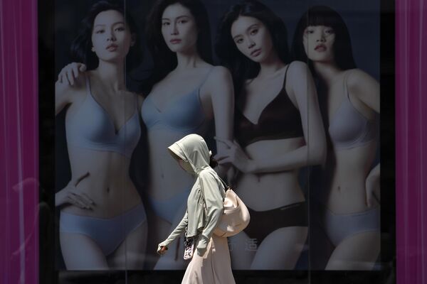 Una mujer con ropa de protección solar camina junto a un cartel publicitario de lencería en el exterior de un centro comercial en un día caluroso y soleado en Pekín, China. - Sputnik Mundo