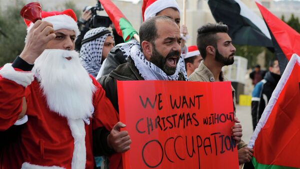 A Palestinian man holds a sign as he stands next to another protester dressed in a Santa Claus costume during a demonstration against the Israeli settlements and demanding for free movement for the Palestinians during Christmas, near a checkpoint in the West Bank city of Bethlehem December 23, 2014 - Sputnik Mundo