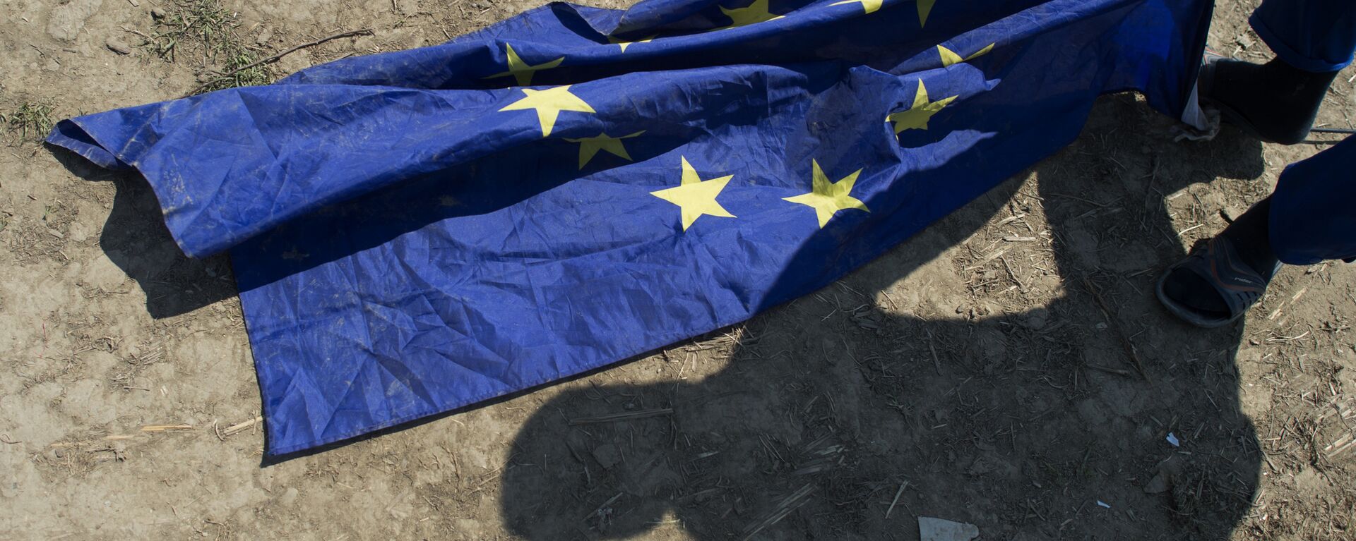 Un migrante de Afganistán toma una bandera de la UE junto a su tienda de campaña en un campamento improvisado para inmigrantes y refugiados cerca de la aldea de Idomeni, no lejos de la frontera entre Grecia y Macedonia, el 1 de mayo de 2016. - Sputnik Mundo, 1920, 15.10.2023