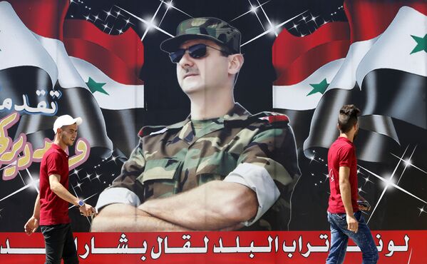 Dos jóvenes pasan por delante de un cartel con la fotografía del presidente sirio, Bashar Asad, en el centro de Damasco. - Sputnik Mundo