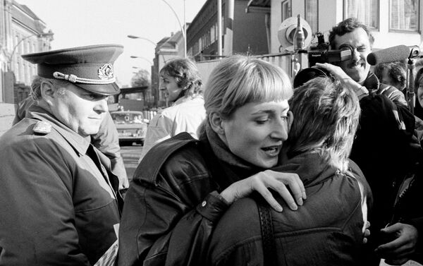 Ciudadanos de Berlín oriental y occidental se abrazan tras la caída del muro mientras un soldado fronterizo los observa - Sputnik Mundo